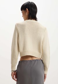 Maglione lavorato a maglia di colore crema, corto, con orlo a coste e maniche lunghe, caratterizzato da una trama testurizzata. Indossato sopra una gonna grigia.