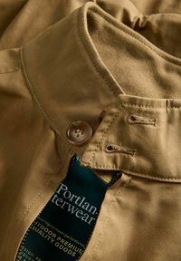 Košeľa béžovej farby s golierom, zapínaním na gombíky a zelenou štítkom vo vnútri, vyrobená z textúrovanej bavlnenej látky s viditeľnými švami.