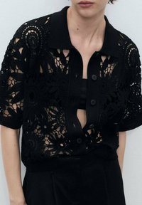 Black lace shirt with short sleeves, featuring intricate floral patterns, open design, and a front button closure. Layered over a black top.