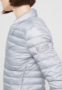 Ljusgrå pufferjacka med horisontella sömmar, slät textur och dragkedja framtill. Har en badge-accent på vänster ärm.