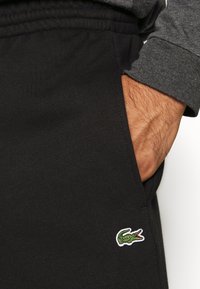 Černé tepláky vyrobené z měkkého materiálu mají elastický pas a boční kapsu, s zeleno-bílým krokodýlem Lacoste poblíž kapsy.