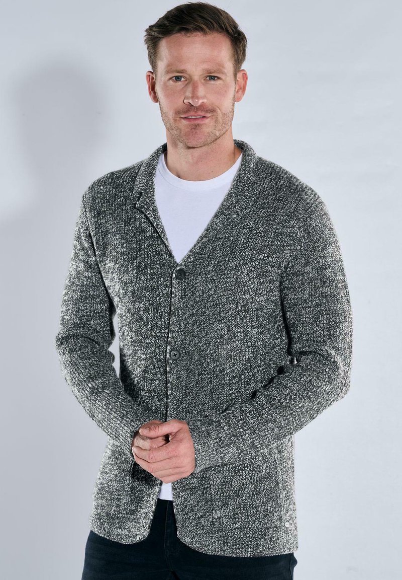 Grau gestrickte Jacke mit strukturiertem Muster, Knopfverschluss und Schalkragen. Über einem weißen T-Shirt getragen, kombiniert mit dunklen Hosen.