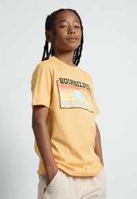 Camiseta de algodón amarilla con un estampado gráfico que presenta un atardecer, palmeras y el texto "Quiksilver" en letras negritas y negras. Mangas cortas.