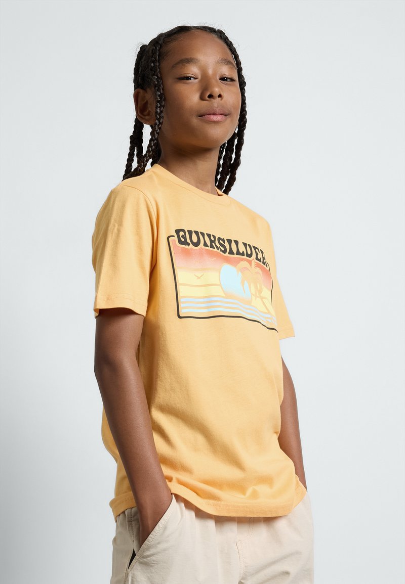 Camiseta de algodón amarilla con un estampado gráfico que presenta un atardecer, palmeras y el texto "Quiksilver" en letras negritas y negras. Mangas cortas.