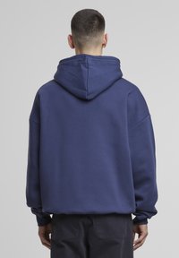 Marineblå hettegenser med stor hette, løs passform, ribbede mansjetter og en myk tekstur. Har dropped shoulders og en klassisk stil.