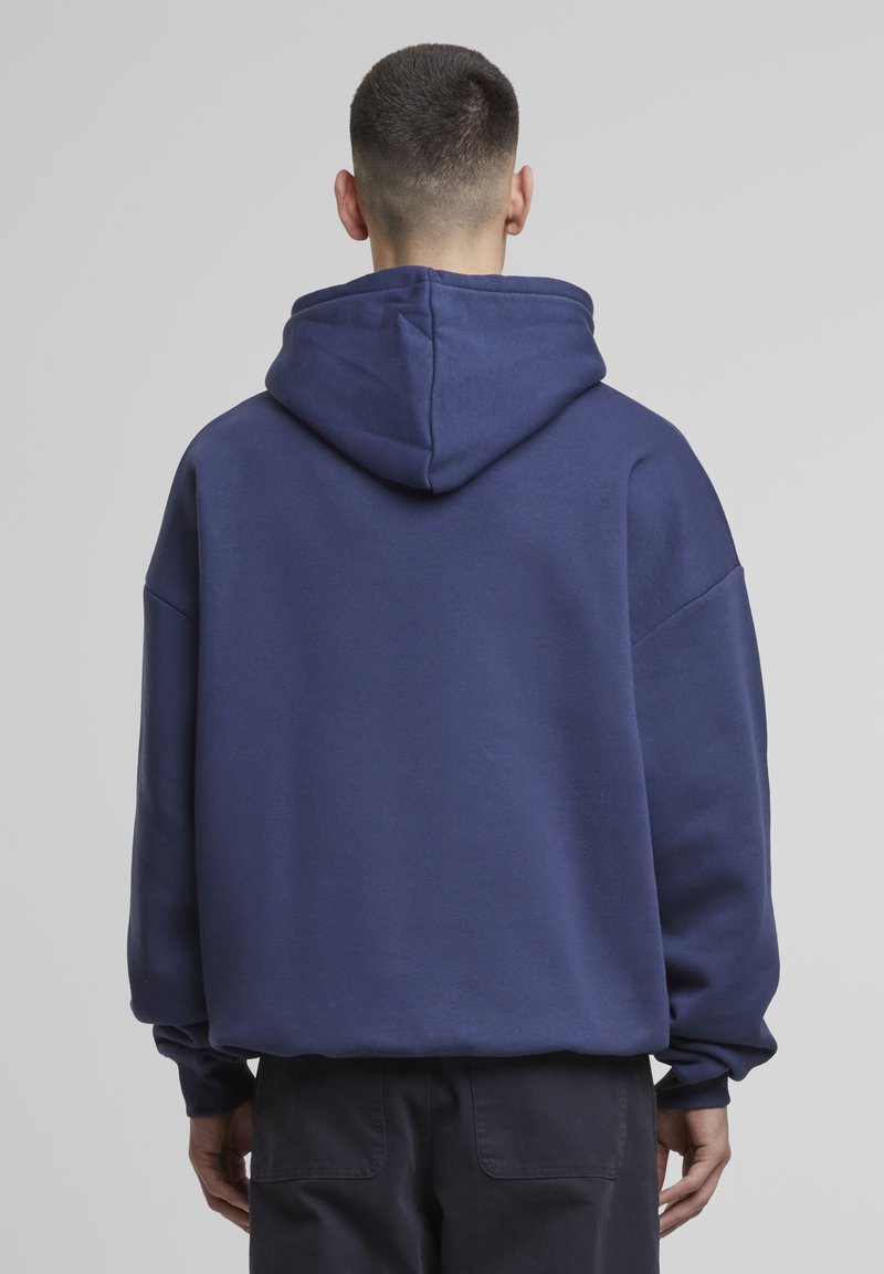 Marineblå hettegenser med stor hette, løs passform, ribbede mansjetter og en myk tekstur. Har dropped shoulders og en klassisk stil.