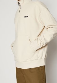 Sudadera de forro polar color crema con cuello de media cremallera, puños acanalados y un bolsillo frontal. Cuenta con un pequeño parche de logo negro en el pecho.