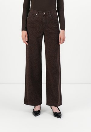 Personne portant un pantalon large marron foncé avec un subtil imprimé léopard, un haut à manches longues noir et des talons pointus noirs, debout sur fond blanc.