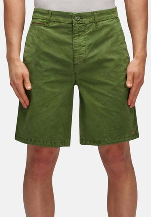 Uomo che indossa pantaloncini casual verde oliva fino al ginocchio con chiusura con bottone e cerniera, in piedi con le mani rilassate accanto alle gambe.