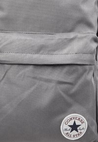 Poche de sac à dos en tissu gris avec des coutures visibles et un patch rond blanc avec le logo Converse All Star en bas à droite.