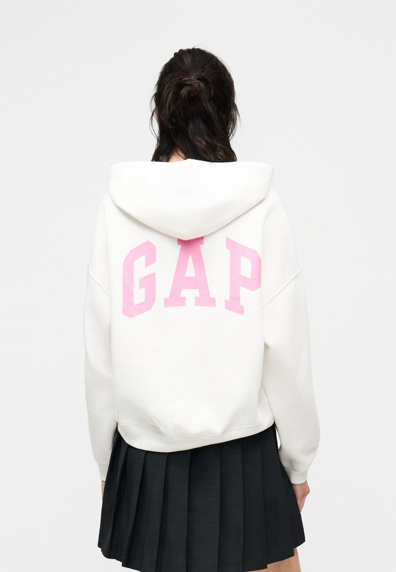Personne aux cheveux foncés portant un sweat à capuche blanc avec de grandes lettres roses "GAP" au dos et une jupe plissée noire, de dos.