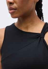 Κοντινό πλάνο ενός ατόμου που φοράει μαύρο τοπ χωρίς μανίκια με λεπτομέρεια δεμένης τιράντας στον δεξιό ώμο και πλεξούδες στα μαλλιά.