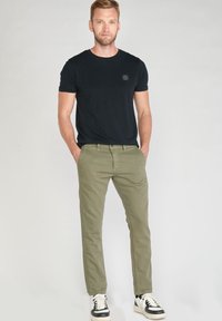 T-shirt noir en coton avec un col rond, associé à un pantalon en coton vert olive. La tenue est complétée par des baskets noires et blanches.