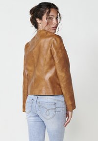Giacca di pelle marrone con un design aderente e una texture liscia, caratterizzata da un alto colletto e dettagli di cucitura semplici. Abbinata a jeans azzurri chiari.