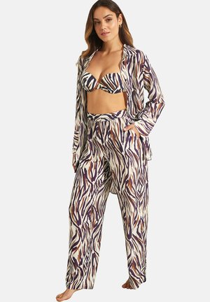 Femme portant un soutien-gorge assorti à rayures zébrées, une chemise ouverte boutonnée et un pantalon ample dans des tons marron, noir et blanc, debout avec une main dans la poche.
