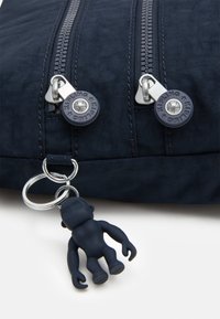 Mørkeblå stoftaske med dobbelt lynlås og sølv hardware. En lille gummimonkey nøglebundt er fastgjort til en ring ved siden af lynlåsene.