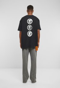 Melna, pārāk liela izmēra T-krekls ar baltu grafisko tekstu aizmugurē, apvienots ar pelēkiem platajiem biksēm un oranžu birku sānos.