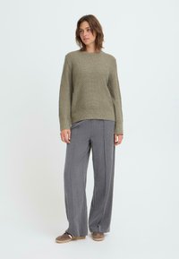 Hellgrüner Strickpullover mit einem strukturierten Muster, kombiniert mit grauen weiten Hosen und braunen Schuhen. Das Modell steht vor einem schlichten Hintergrund.