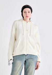 Kremfarget zip-opp hettegenser med stor hette, snor og frontlommer. Kombinert med lyseblå stripete jeans.