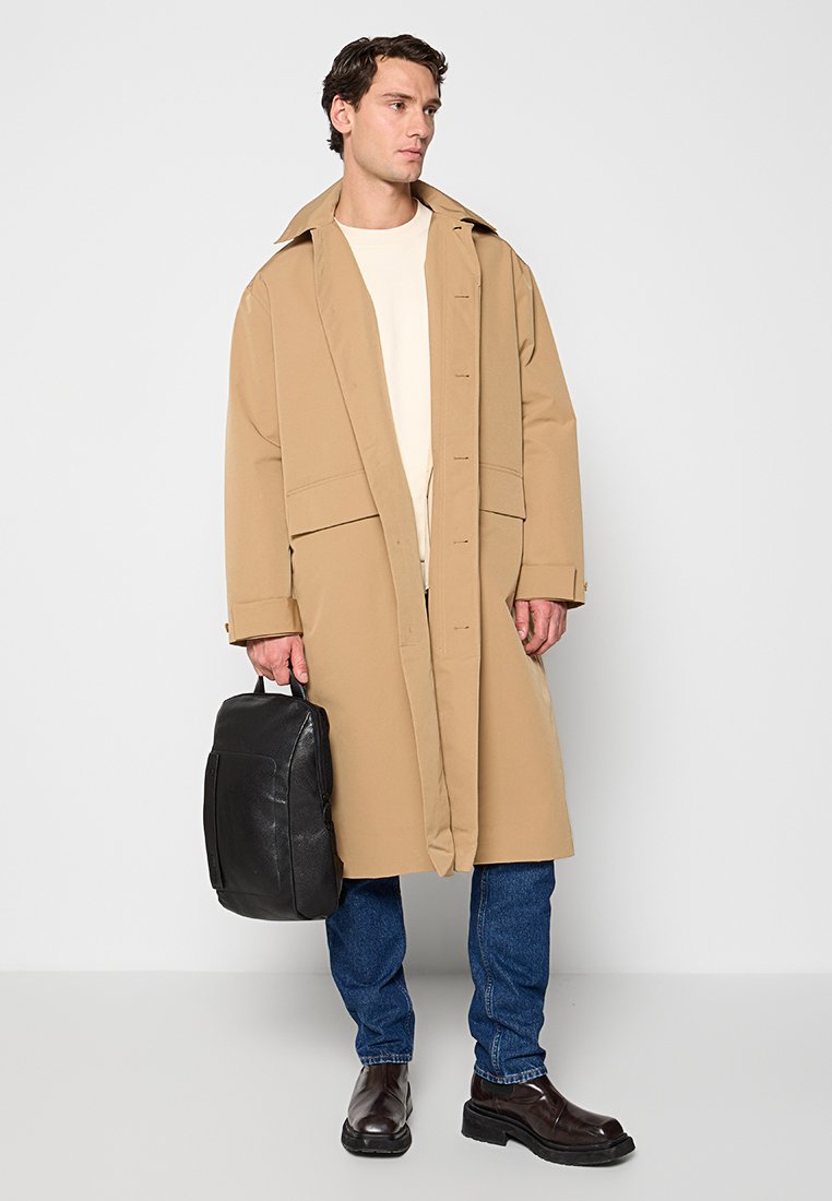 Beige oversized jas met knoopsluiting, grote kraag en voorzakken, gedragen over een lichte trui en spijkerbroek. Zwarte rugzak en bruine laarzen.