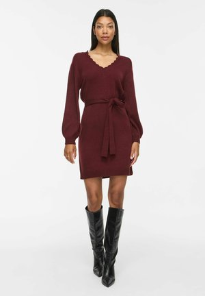 Femme portant une robe en maille bordeaux à manches longues avec un col en V festonné et une ceinture à la taille, associée à des bottes en cuir noires montantes jusqu'aux genoux.