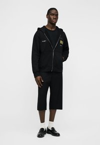 Άνδρας που στέκεται φορώντας μαύρο φούτερ με φερμουάρ, μαύρα σορτς μέχρι το γόνατο, λευκές κάλτσες και μαύρα μοκασίνια, μπροστά σε απλό λευκό φόντο.