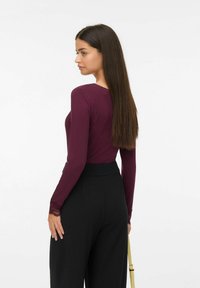 Femme aux longs cheveux noirs portant un haut violet à manches longues avec des poignets en dentelle et un pantalon noir taille haute, tenant un sac à bandoulière beige.