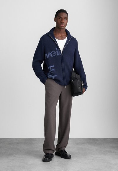 Marinblå zip-up tröja med ribbad krage, prydd med ljusblå text. Matchad med grå byxor och svarta skor, håller en svart väska.
