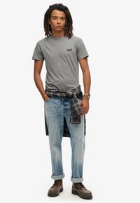 Grijze T-shirt met korte mouwen en een klein logo, lichtblauwe jeans opgerold bij de manchetten, zwart-wit geruit overhemd om de taille geknoopt, bruine laarzen.