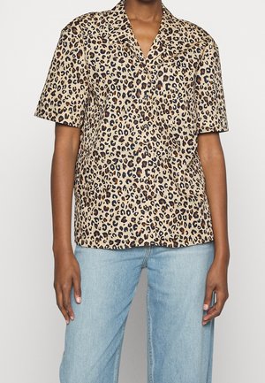 Persona que lleva una camisa beige de manga corta con un estampado de leopardo negro y marrón combinada con vaqueros de mezclilla azul claro.