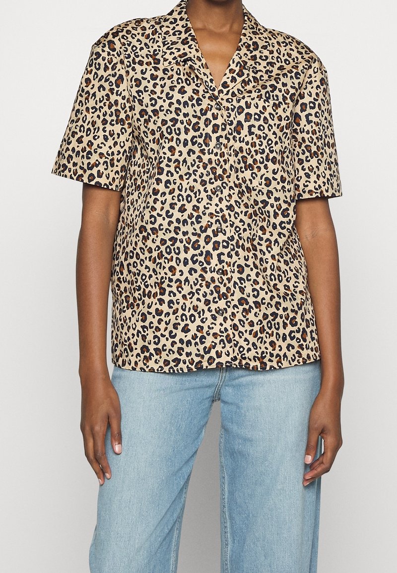 Personne portant une chemise beige à manches courtes avec un motif léopard noir et marron, associée à un jean en denim bleu clair.