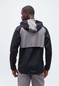 Άνδ ρας με μαύρο και γκρι μπουφάν με κουκούλα και ταιριαστό γκρι παντελόνι, γυρισμένος με την πλάτη προς το μέρος μας, μπροστά σε απλό ανοιχτό φόντο.