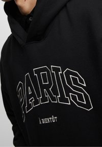 Sudadera negra con gran texto blanco "PARIS" en un diseño curvado, y texto más pequeño "à BIENTÔT" debajo. Tejido suave, corte casual y capucha.