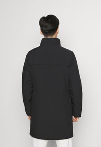 Svart lang frakk med høy krage, glatt stoff og minimalistisk design. Har en rett bakside og detaljer i sidesømmen.