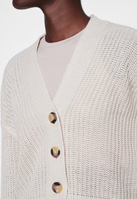 Beige stickad cardigan med V-ringning, prydd med tre stora, runda knappar i guldton. Mönstrad textil och öppen stickning.