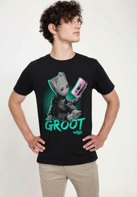 Zwarte katoenen t-shirt met een afbeelding van Groot die een cassettebandje vasthoudt. Het ontwerp bevat groene accenten en gestileerde tekst met de tekst "GROOT."