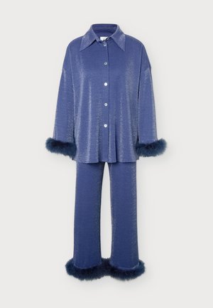 Femme porte une chemise bleue brillante boutonnée et un pantalon large assorti, tous deux ornés de poignets en plumes bleu foncé, avec des talons ouverts blancs.