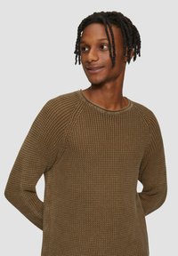 Pull en maille marron avec un motif texturé, des manches longues et un col rond. Le tissu a un aspect côtelé et une coupe décontractée.