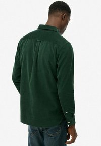 Hombre vestido con una camisa de manga larga verde oscuro y jeans azules, de espaldas contra un fondo blanco liso.
