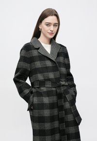 Casaco xadrez preto e cinza com colarinho tipo echarpe, cintura com cinto e bolsos grandes. Tecido macio com design estruturado.