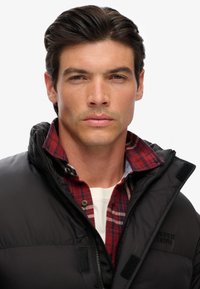 Chaqueta de plumas negra con un cuello alto, usada sobre una camisa de cuadros roja y azul. Presenta cierres de cremallera y velcro, con un logo en el pecho.