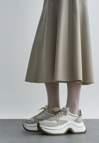 Beežid tossud tekstuurse kangaga, ümarate valgete tallade ja oliiviroheliste aktsentidega. Disainis on paelad ja dekoratiivne logo.