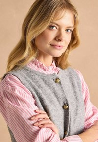 Graham grüne Weste mit goldenen Knöpfen, die über einer rosa und weißen gestreiften Bluse mit Rüschensaum und langen Ärmeln getragen wird.