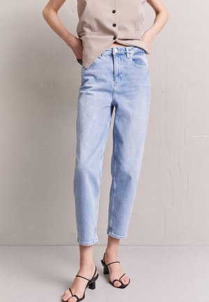 Persoon draagt lichtblauwe jeans met hoge taille en een beige mouwloze top, staat met handen in de zakken en draagt zwarte sandalen met hak.