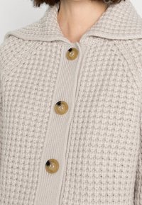 Svetr s waffle pleteninou v světle béžové barvě se širokým límcem, velkými hnědými knoflíky, vroubkovaným předním zapínáním a texturovanou tkaninou.
