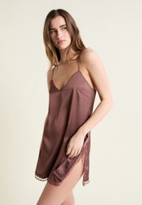 Joven con el cabello largo y castaño, vistiendo un vestido tipo slip de satén color malva con ribete de encaje, de pie frente a un fondo claro y liso.