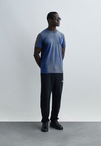 Άνδρας που φοράει μαύρα γυαλιά ηλίου, μπλε μπλουζάκι με βαθμιδωτό χρώμα και λογότυπο "M", μαύρο παντελόνι τζόγκερ και μαύρα αθλητικά παπούτσια, στέκεται με τα χέρια πίσω από την πλάτη μπροστά σε απλό φόντο.