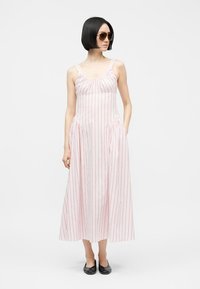 Žena s krátkymi čiernymi vlasmi, ktorá nosí slnečné okuliare a biele šaty bez rukávov s vertikálnymi červenými pruhmi a čierne topánky na nízkom opätku.