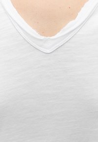 Gros plan d'un col de chemise blanche en V avec des coutures visibles et des bords légèrement effilochés sur un fond de peau claire.