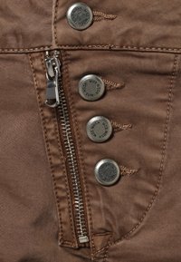 Brown fabric close-up featuring a silver zipper, four metallic snap buttons, and stitched detailing, highlighting a smooth texture.